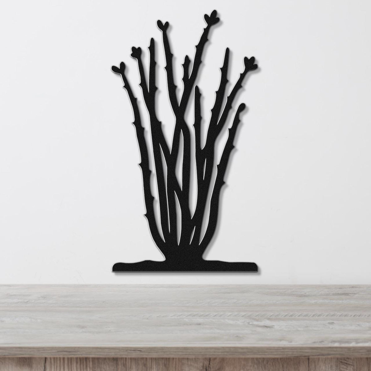Black metal ocotillo wall art with branching cutouts and leaf shapes, mounted on a white wall above a light wood surface.