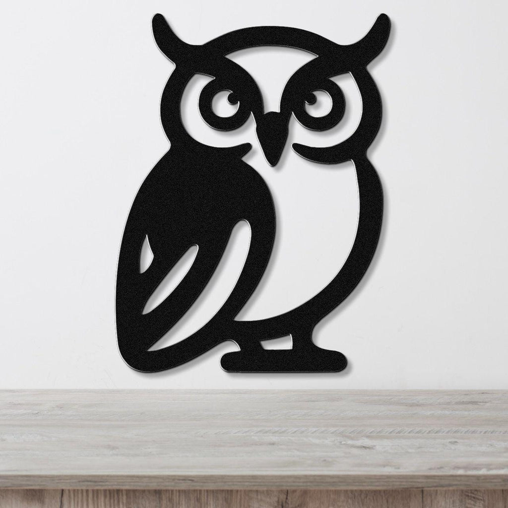 Black metal owl wall art with cutout eyes and ear tufts, mounted on a white wall above a wooden surface.