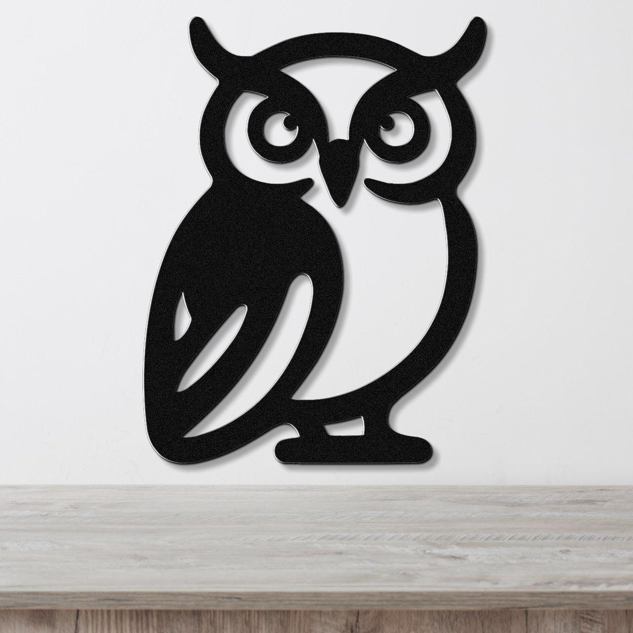 Black metal owl wall art with cutout eyes and ear tufts, mounted on a white wall above a wooden surface.