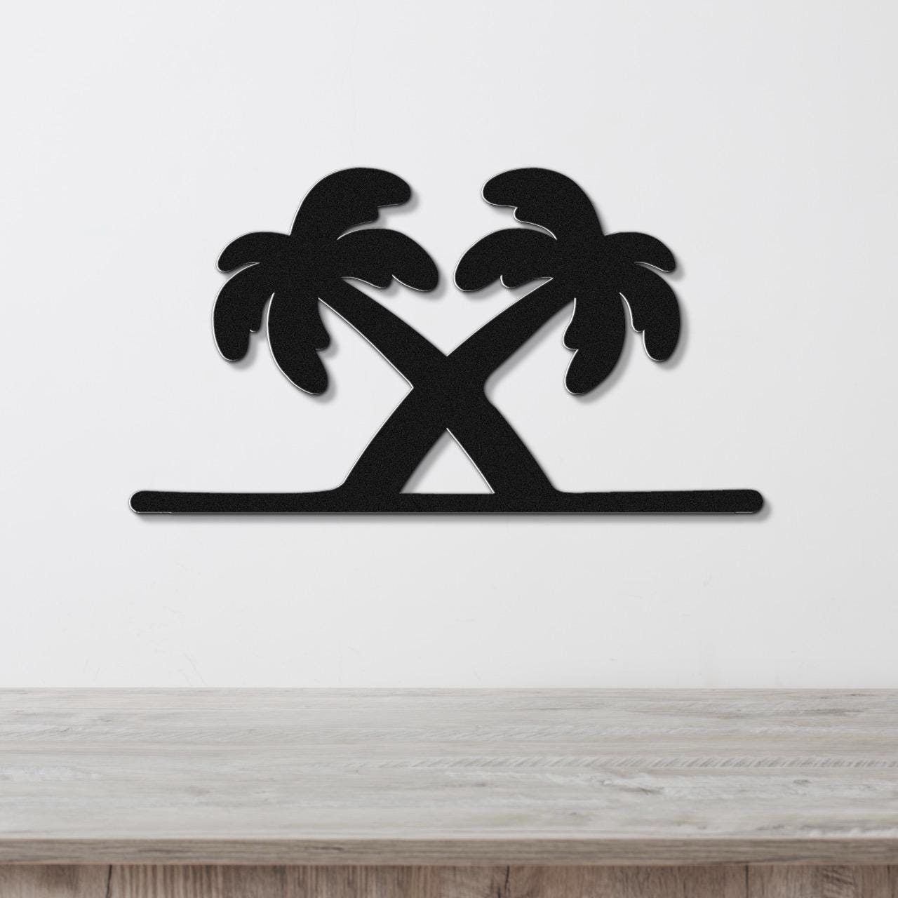 Black metal wall art featuring a silhouette of two crossed palm trees with leafy fronds, mounted on a white wall above a light wood surface.