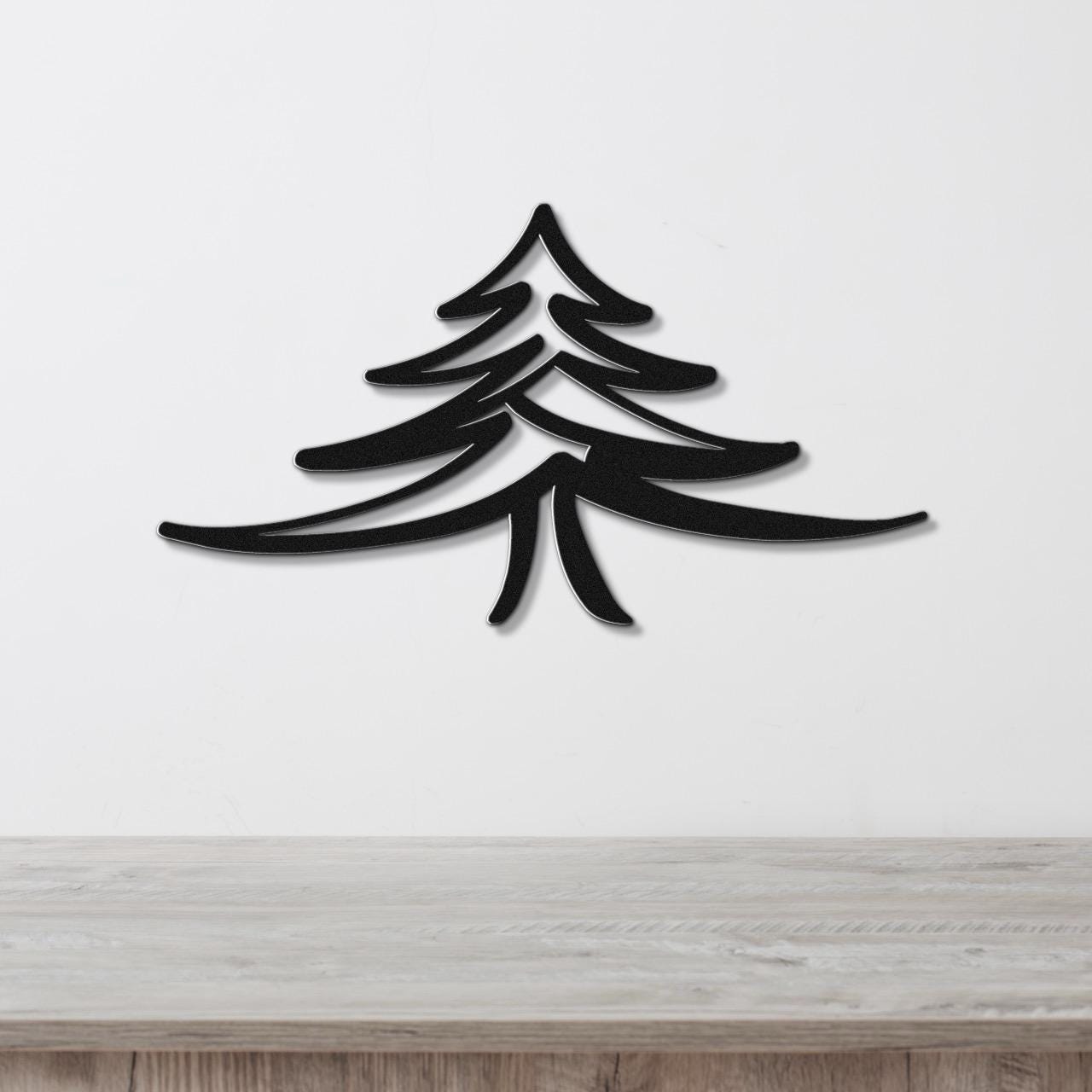 Black metal pine tree wall art with layered branch cutouts, mounted on a white wall above a light wood surface.