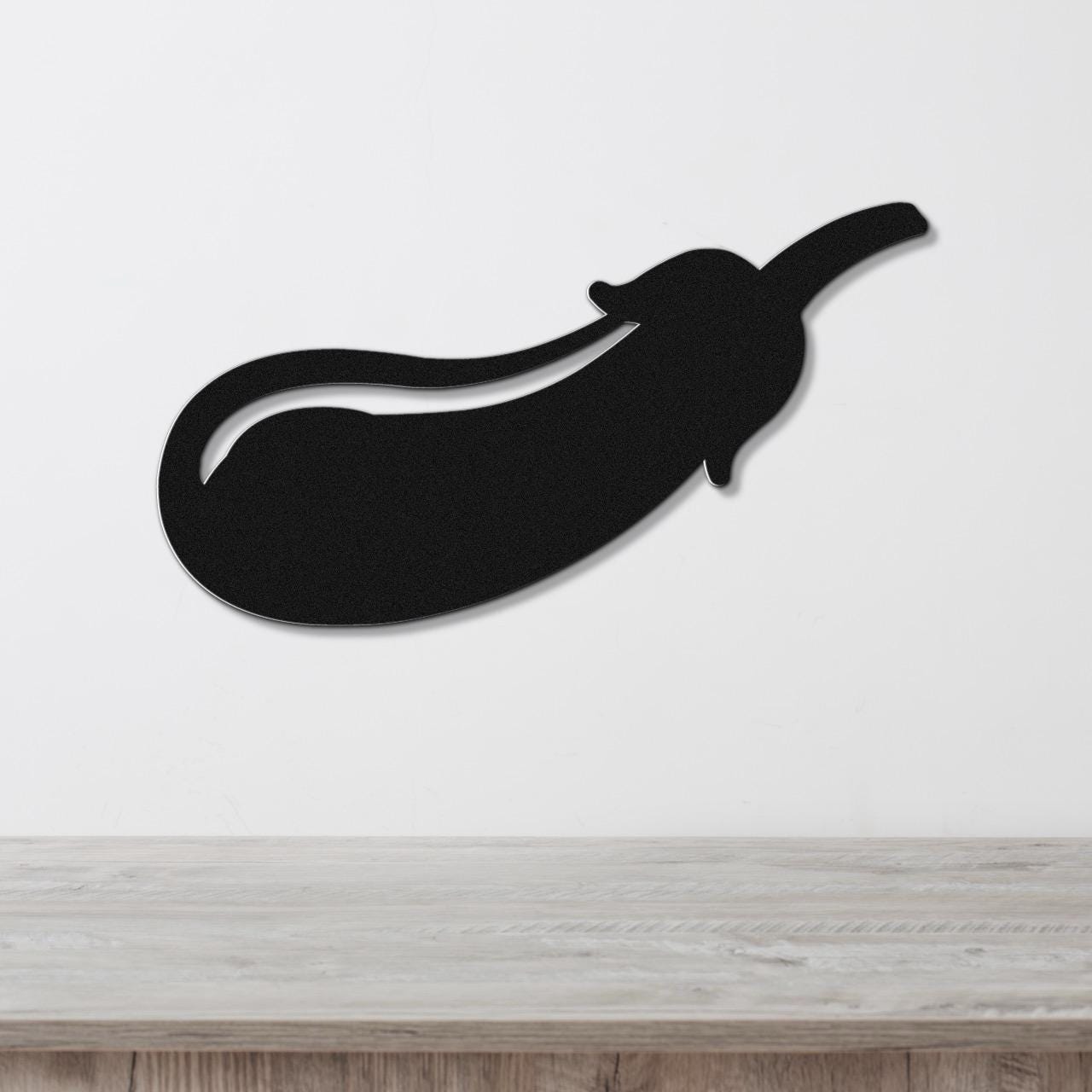 Black metal eggplant wall art with a cutout stem and curved body, mounted on a white wall above a light wood surface.