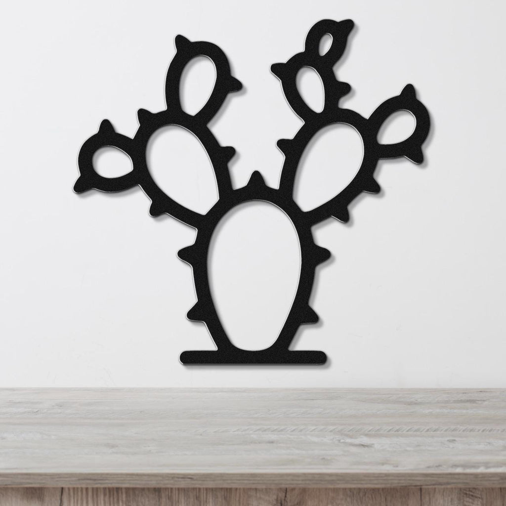 Black metal wall art cutout of a prickly pear cactus with multiple pads and spines, mounted on a white wall above a light wood surface.