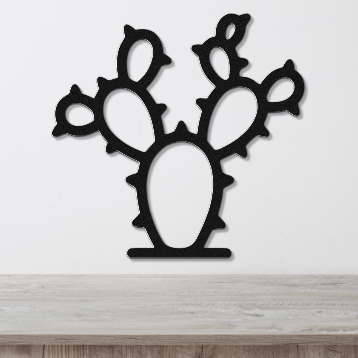 Black metal wall art cutout of a prickly pear cactus with multiple pads and spines, mounted on a white wall above a light wood surface.