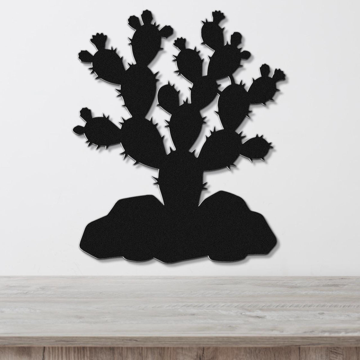 Black metal wall art cutout of a prickly pear cactus with multiple pads and spines, mounted on a white wall above a light wood surface.