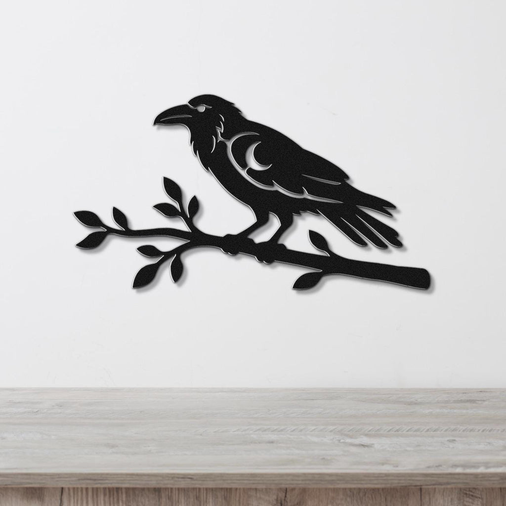 Black metal wall art of a raven perched on a branch with a crescent moon cutout, mounted on a white wall above a wooden surface.