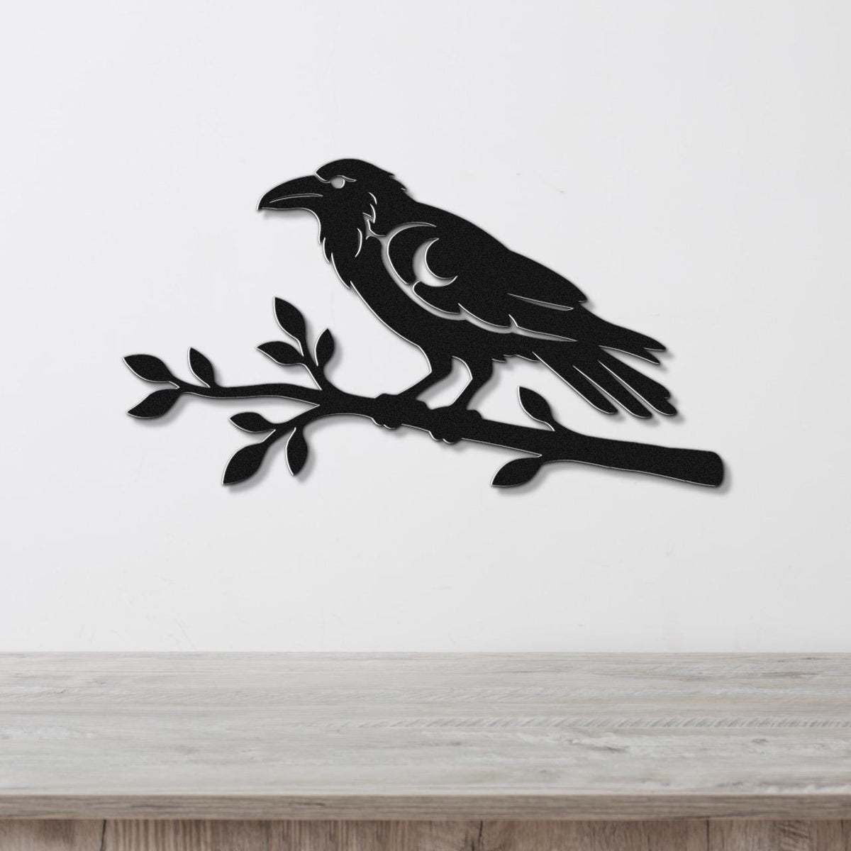 Black metal wall art of a raven perched on a branch with a crescent moon cutout, mounted on a white wall above a wooden surface.