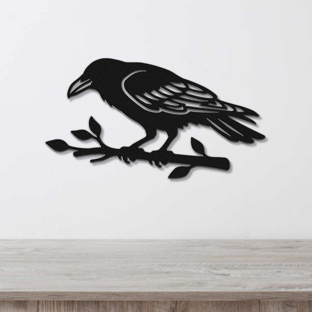 Black metal raven wall art with detailed feather cutouts, perched on a branch, mounted on a white wall above a wooden surface.