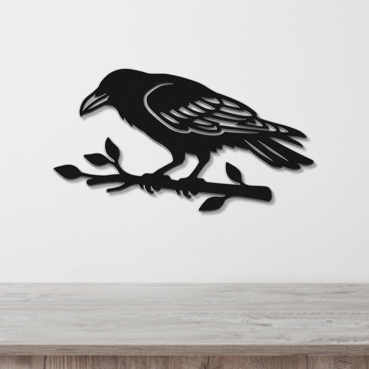 Black metal raven wall art with detailed feather cutouts, perched on a branch, mounted on a white wall above a wooden surface.