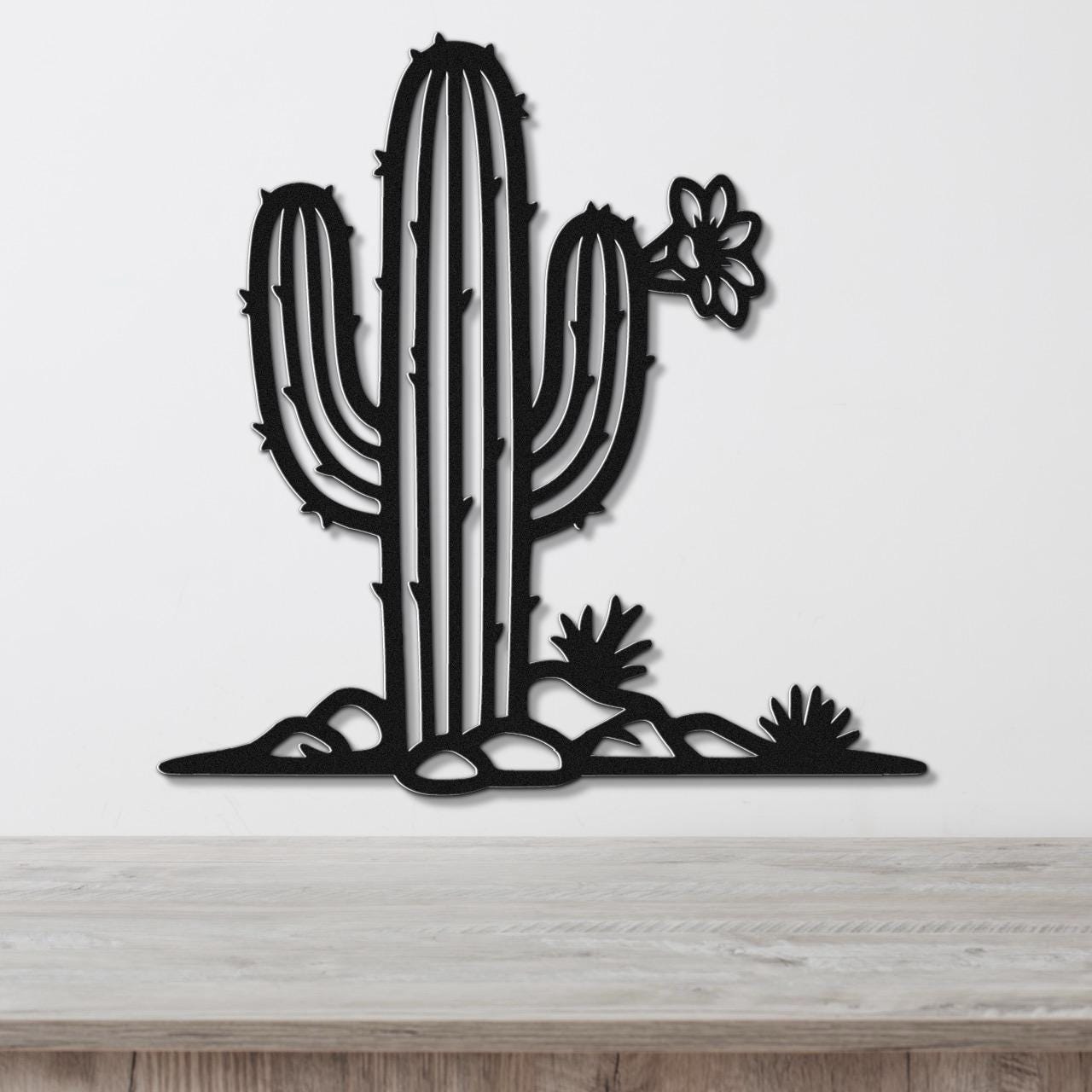 Black metal wall art cutout of a saguaro cactus with flower and smaller cacti, mounted on a white wall above a wooden surface.