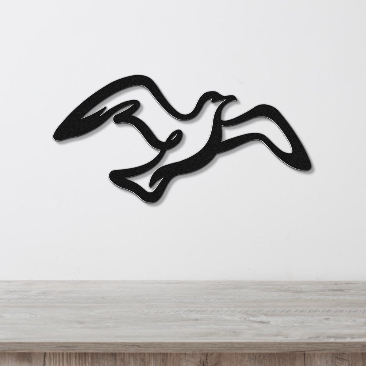 Black metal seagull wall art with flowing wing cutouts, mounted on a white wall above a light wood surface.