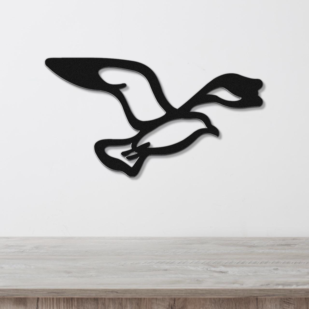 Black metal seagull wall art with wing and tail cutouts, mounted on a white wall above a light wood surface.