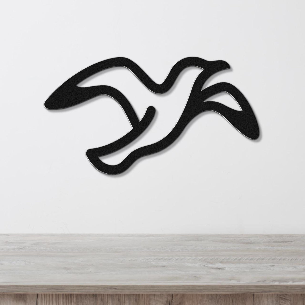 Black metal seagull wall art with flowing motion lines, mounted on a white wall above a light wood surface.