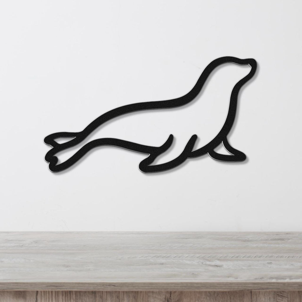 Black metal sea lion wall art in a sleek silhouette cutout style, mounted on a white wall above a light wood surface.