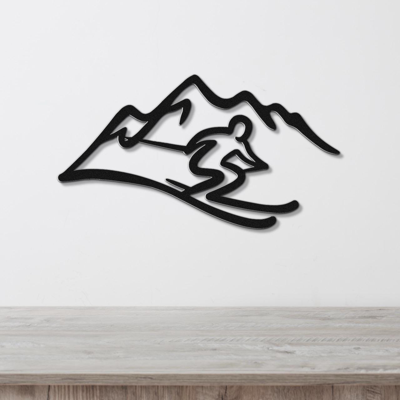 Black metal wall art featuring a skier silhouette in motion against a mountain range, mounted on a white wall above a wooden surface.