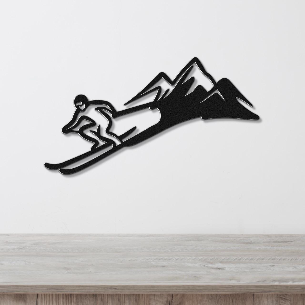 Black metal wall art featuring a skier silhouette in motion with mountain peaks, mounted on a white wall above a wooden surface.