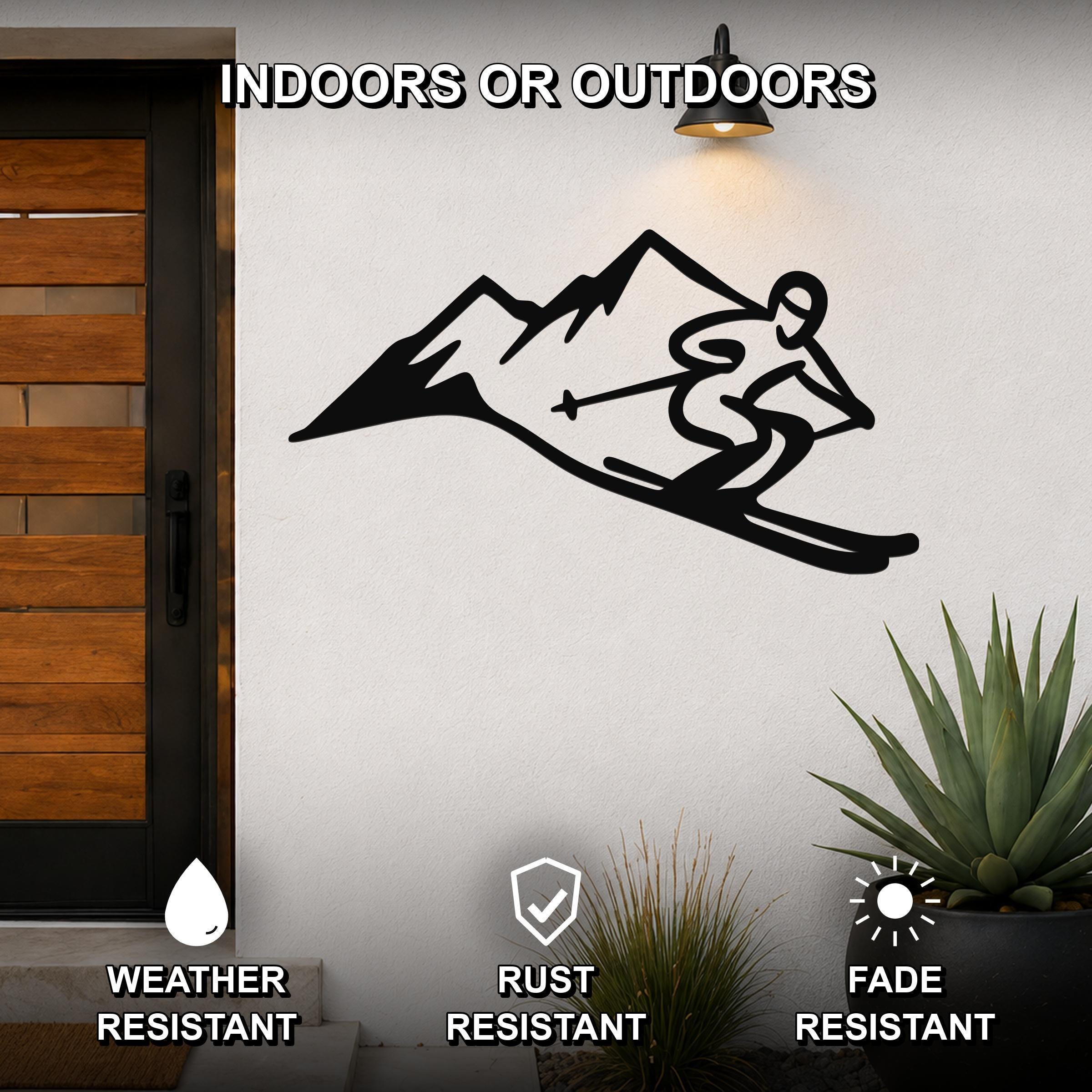 Black metal wall art featuring a skier silhouette in motion above a mountain range, mounted on an outdoor wall beside a wooden door and potted plants.