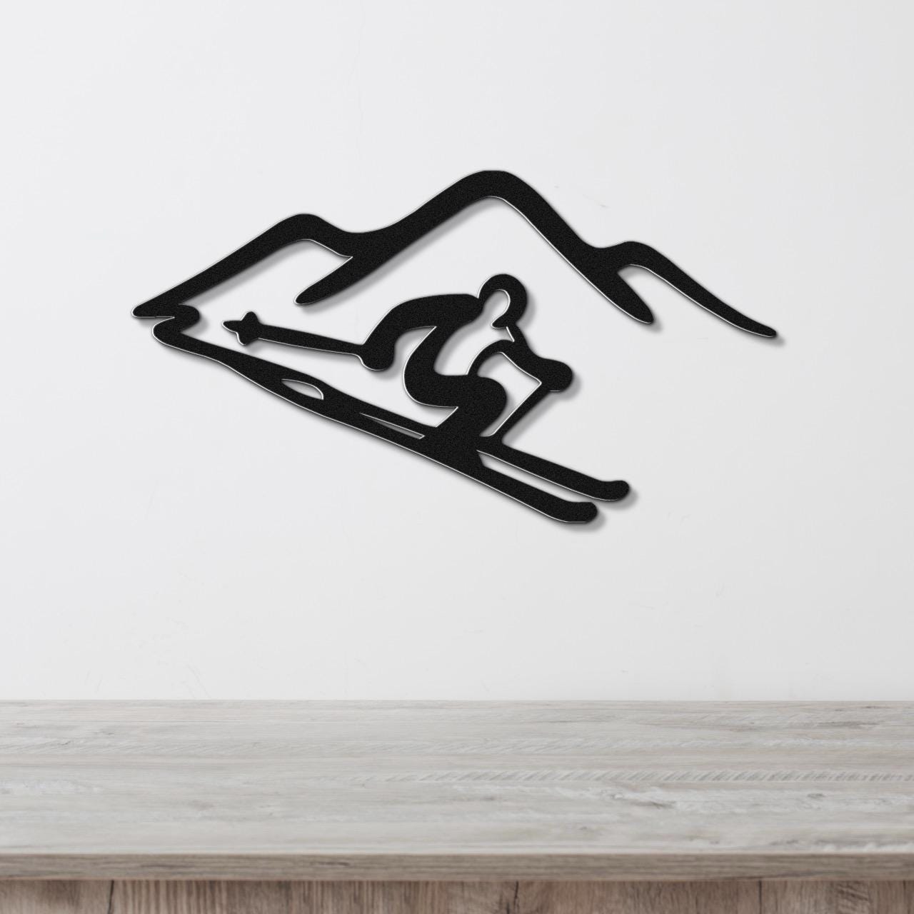 Black metal wall art featuring a skier silhouette and mountain cutout, mounted on a white wall above a wooden surface.