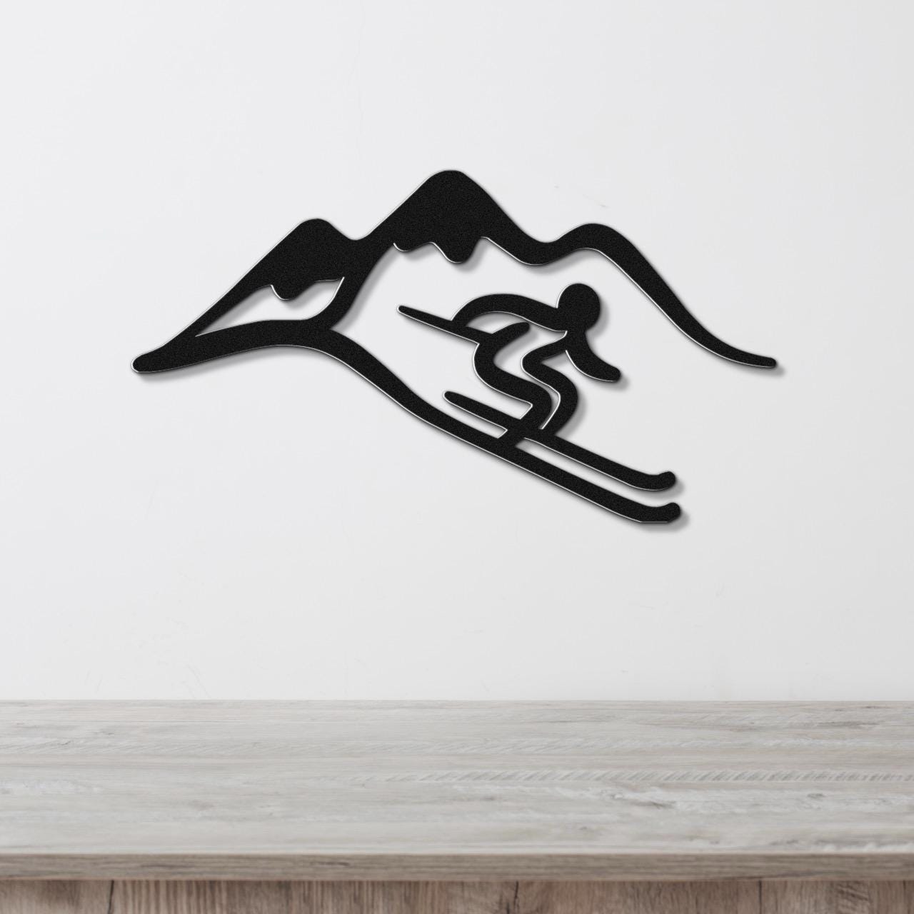 Black metal wall art featuring a skier silhouette in motion against a mountain range, mounted on a white wall above a light wood surface.