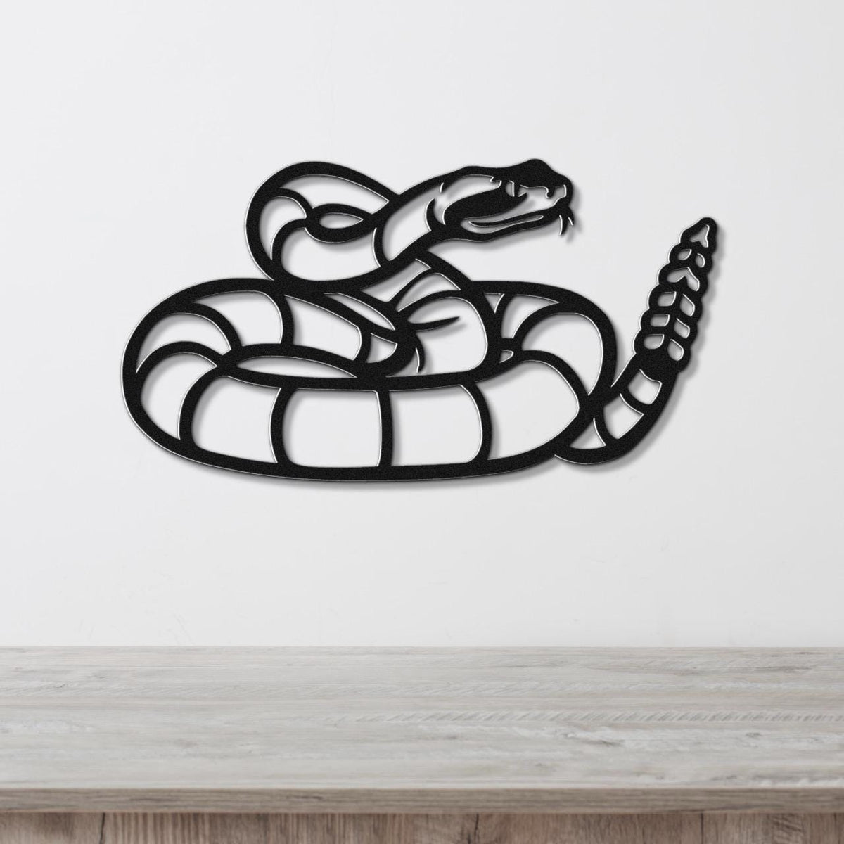 Black metal snake wall art with coiled body and raised head, featuring internal cutout details, mounted on a white wall above a wooden surface.