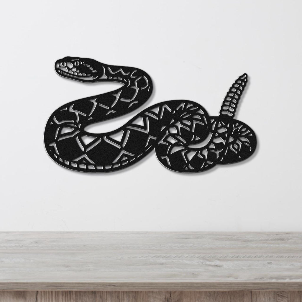 Black metal wall art sculpture of a coiled rattlesnake with detailed internal cutouts, mounted on a white wall above a light wood surface.