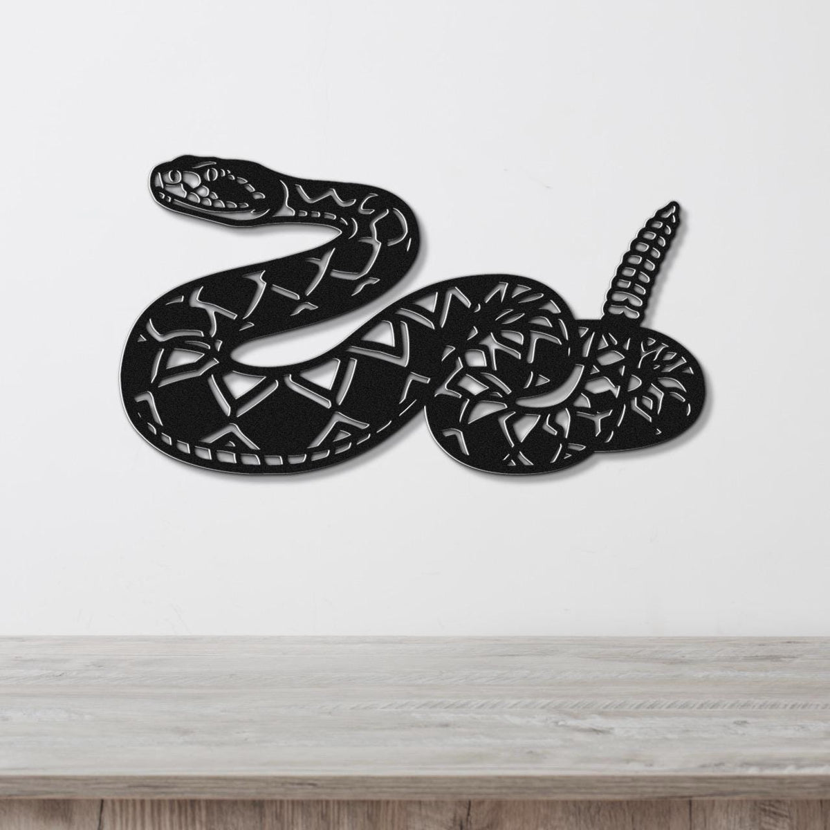 Black metal wall art sculpture of a coiled rattlesnake with detailed internal cutouts, mounted on a white wall above a light wood surface.