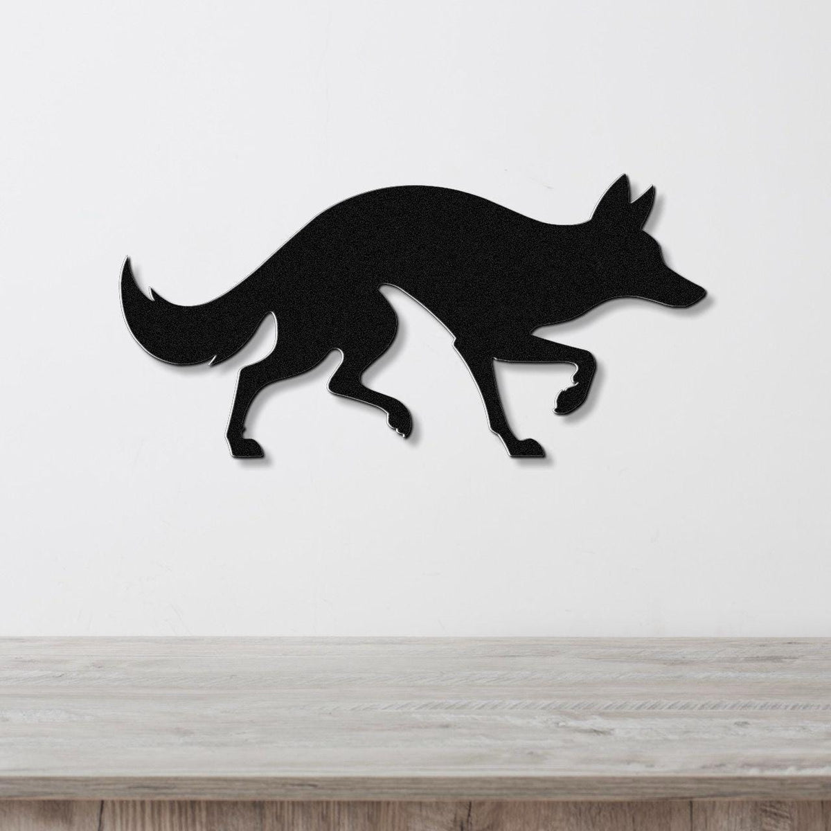 Black metal coyote wall art silhouette with a curved tail and raised front paw, mounted on a white wall above a light wood surface.