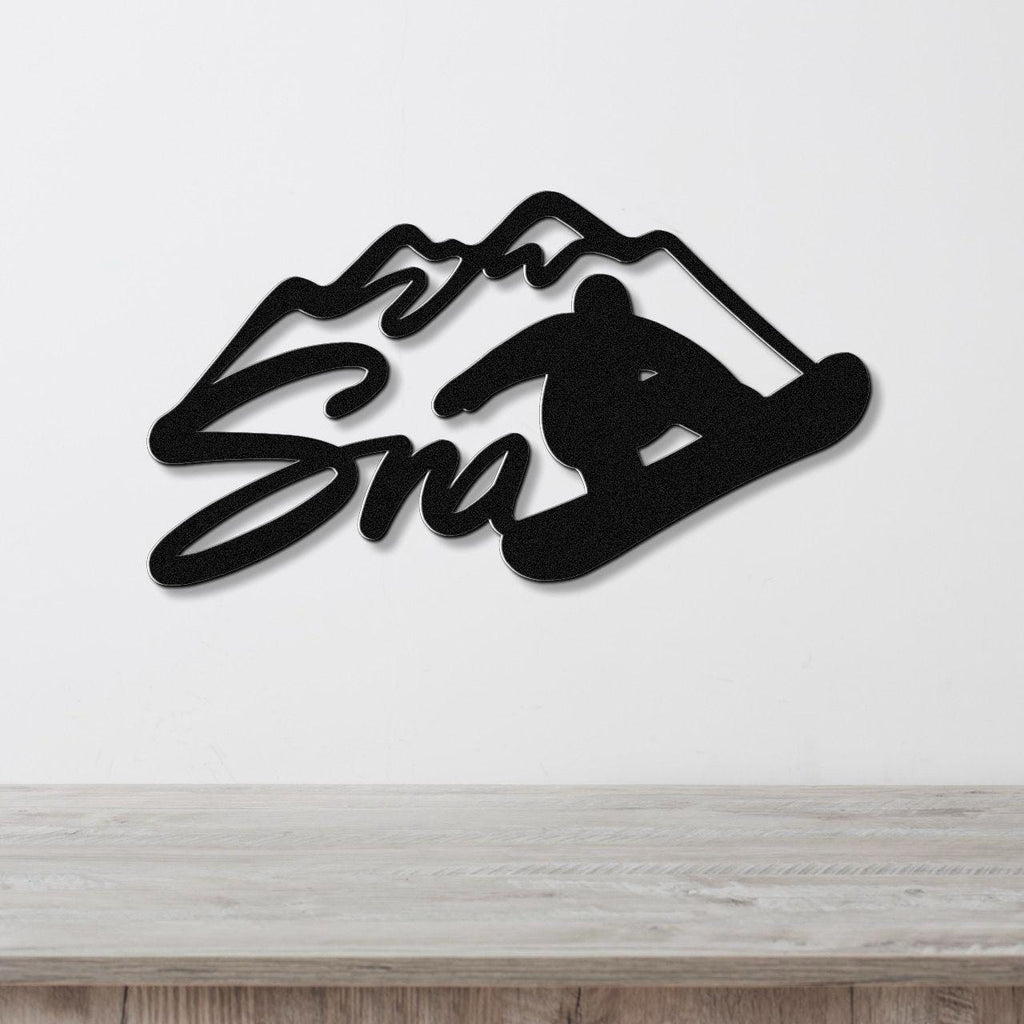 Black metal wall art featuring a snowboarder silhouette and mountain peaks with the word "Snow" in cursive, mounted on a white wall above a wooden surface.