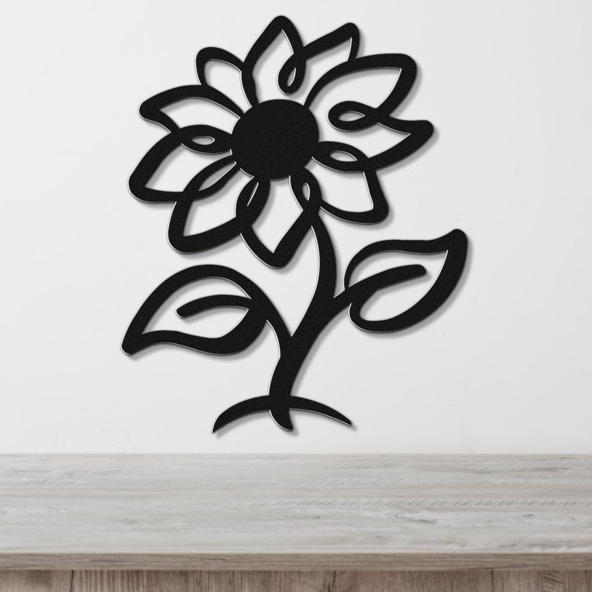 Black metal sunflower wall art with cutout petals and leaves, mounted on a white wall above a light wood surface.