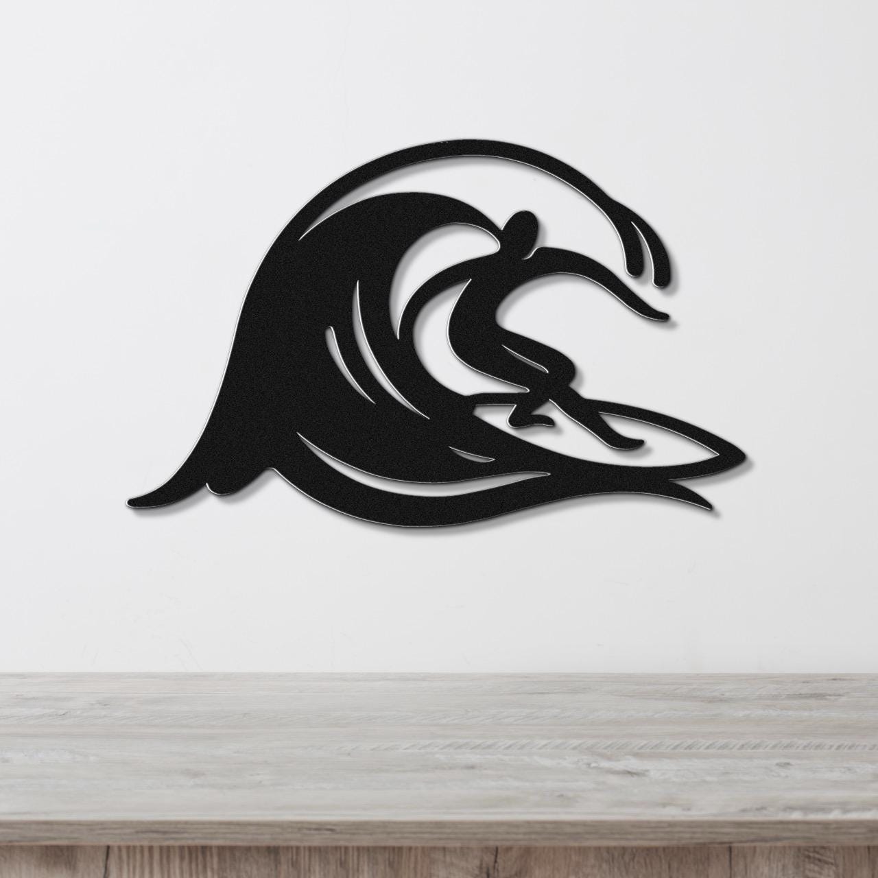 Black metal wall art featuring a surfer silhouette riding a flowing wave, mounted on a white wall above a wooden surface.