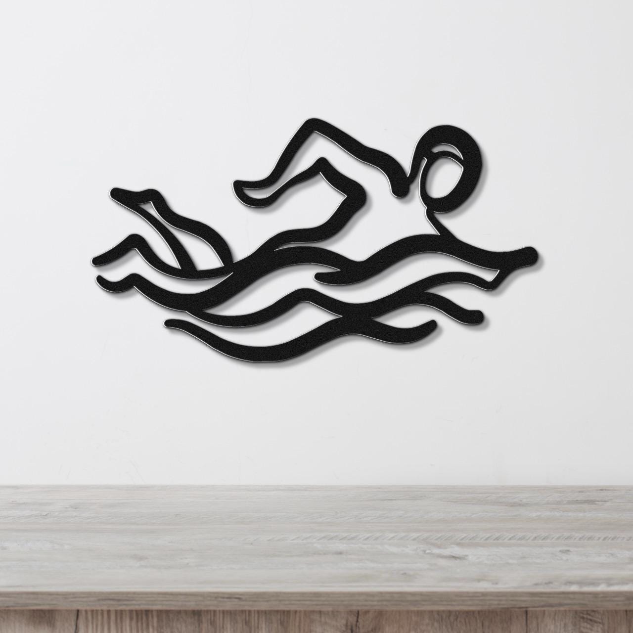 Black metal wall art depicting a swimmer in motion with wavy water lines, mounted on a white wall above a wooden surface.