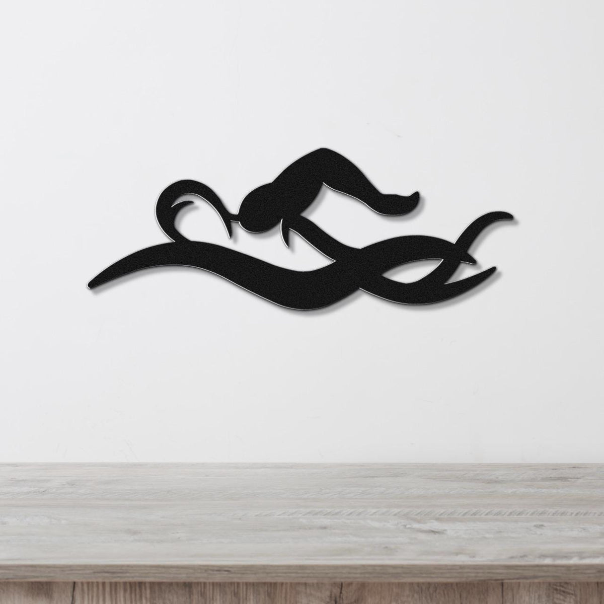 Black metal wall art of a swimmer in motion, with fluid cutout lines, mounted on a white wall above a wooden surface.