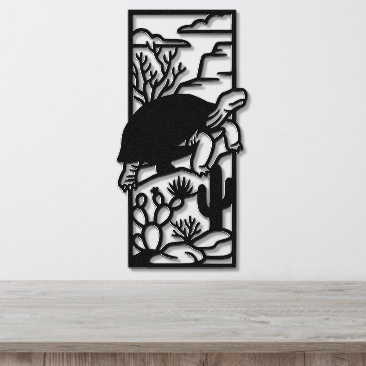 Black metal wall art featuring a tortoise silhouette climbing over cacti and desert plants, mounted on a white wall above a wooden surface.