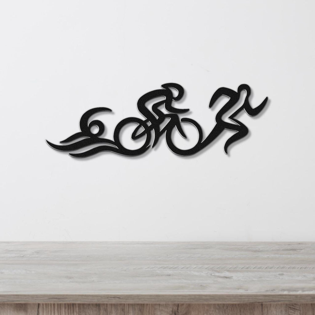 Black metal triathlon wall art with flowing cutout design of swim, bike, and run figures, mounted on a white wall above a wooden surface.