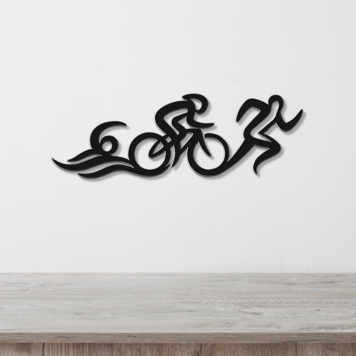 Black metal triathlon wall art with flowing cutout design of swim, bike, and run figures, mounted on a white wall above a wooden surface.