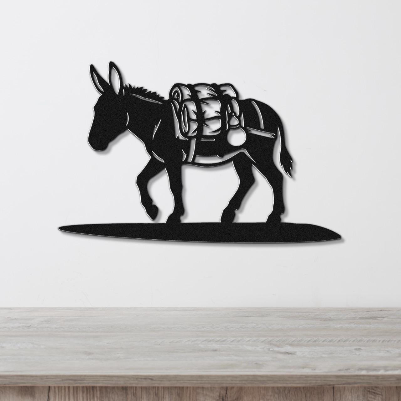 Black metal burro wall art silhouette with saddle and pack details, mounted on a white wall above a wooden surface.