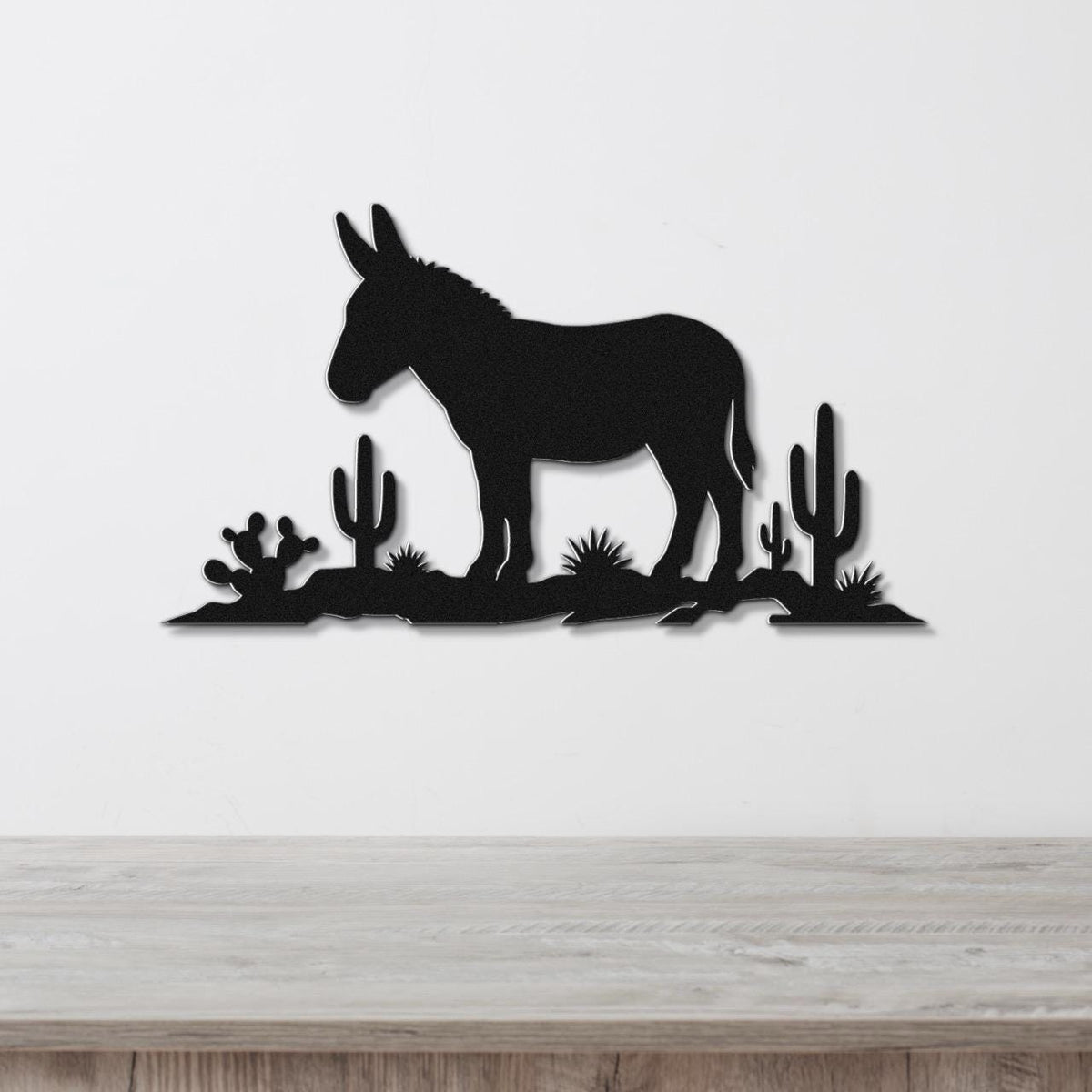 Black metal wall sculpture of a burro silhouette with cacti, mounted on a white wall above a wooden surface.