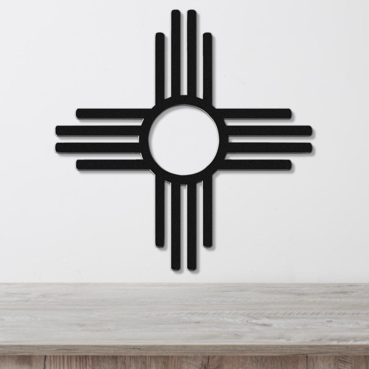 Black metal Zia Sun symbol wall art with a bold circular cutout and radiating rays, mounted on a white wall above a light wood surface.