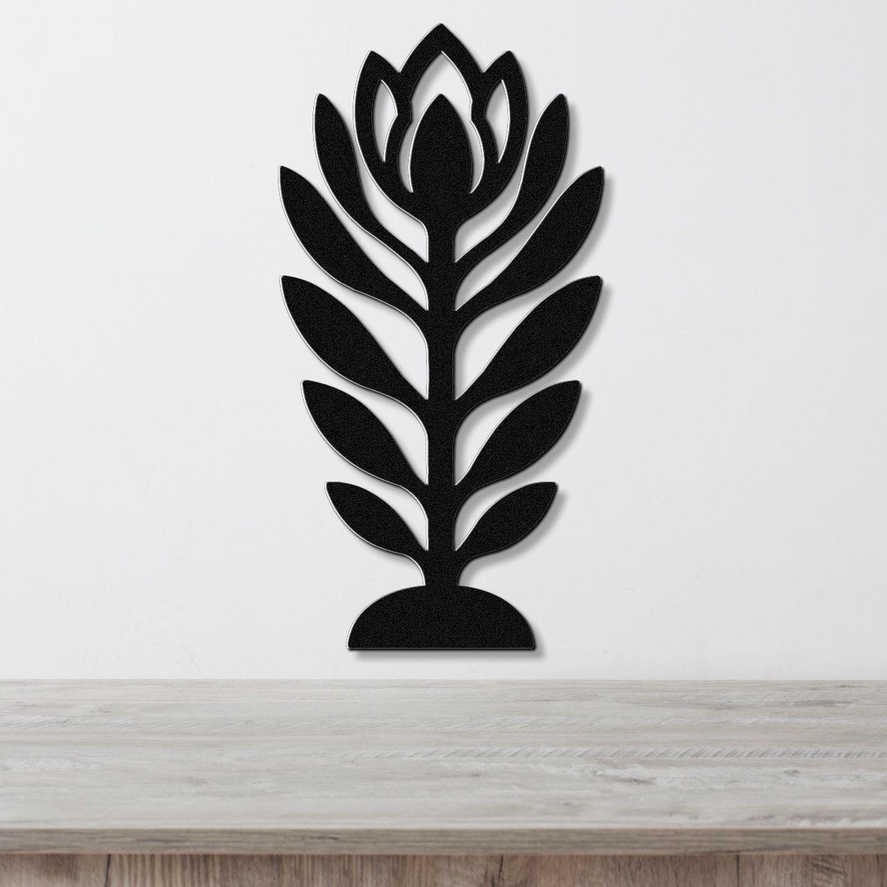 Black metal wall art in a stylized wildflower silhouette with layered leaf and bloom cutouts, mounted on a white wall above a light wood surface.