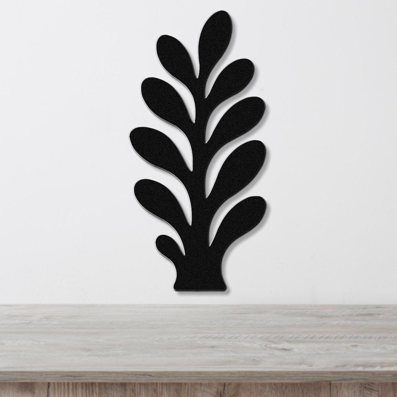 Black metal wall art in a leafy sprig silhouette with cutout details, mounted on a white wall above a light wood surface.