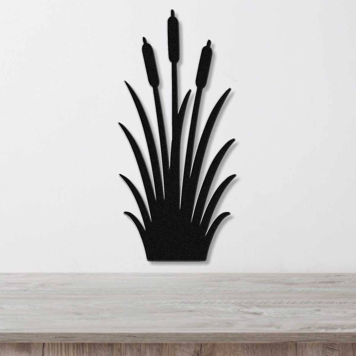 Black metal cattail wall art with a silhouette design of tall reeds and seed heads, mounted on a white wall above a light wood surface.