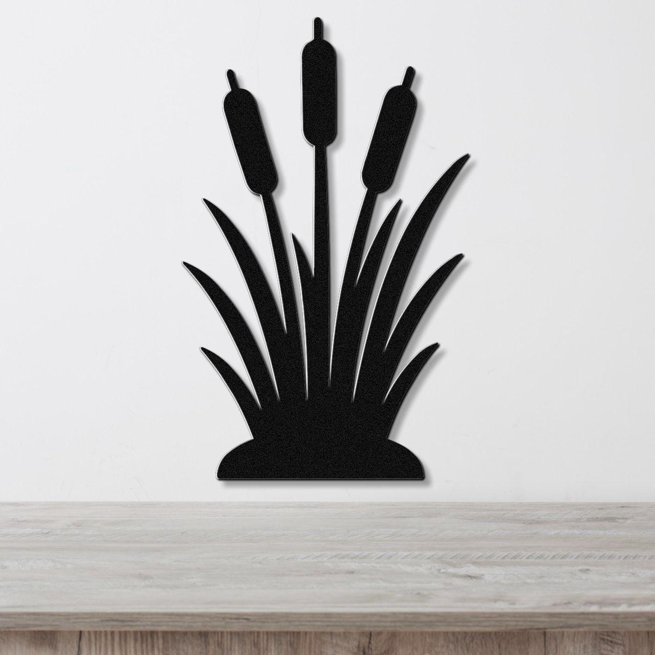 Black metal wall art silhouette of cattails with three seed heads and long leaves, mounted on a white wall above a light wood surface.