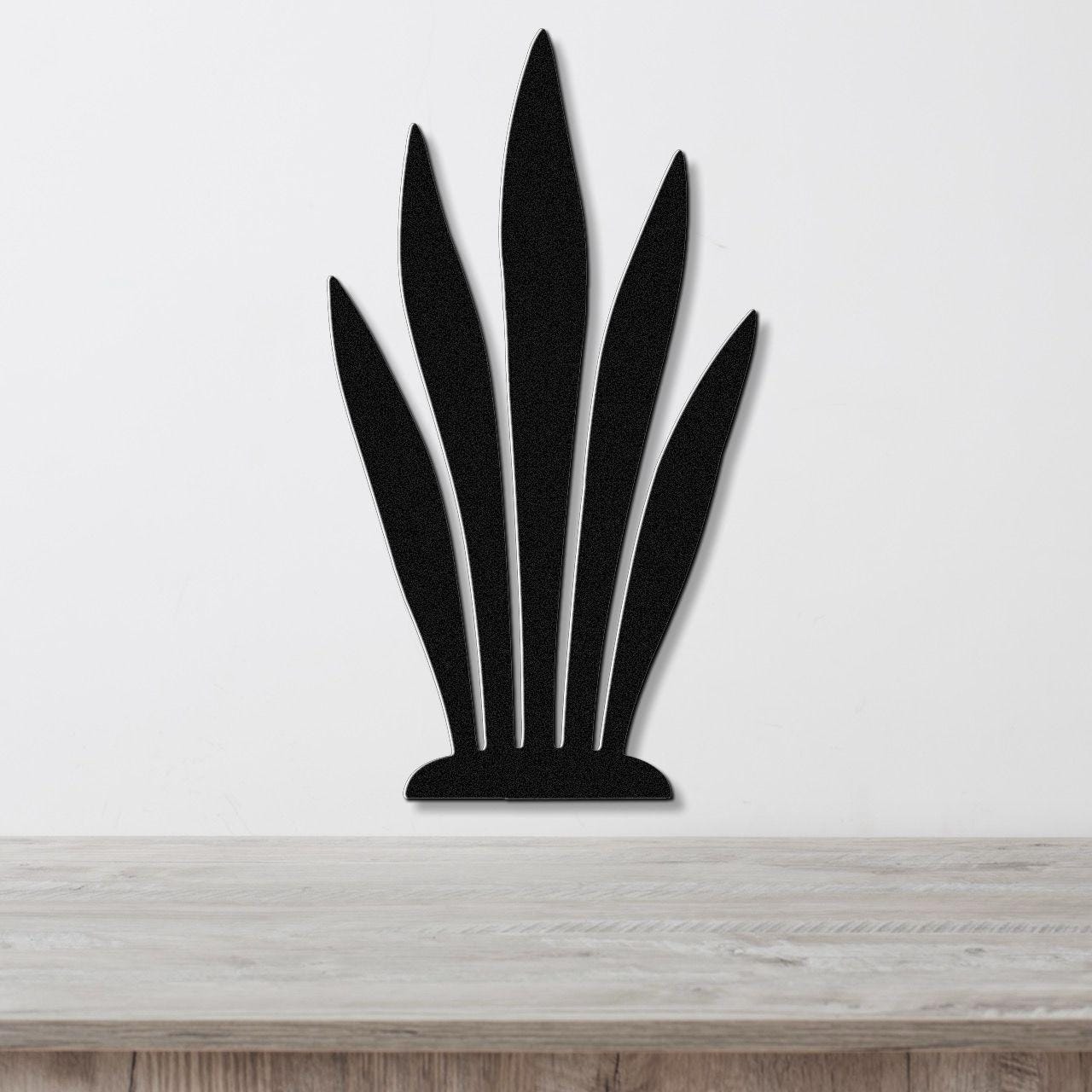 Black metal wall art in a snake plant silhouette with five tall, pointed leaves, mounted on a white wall above a light wood surface.