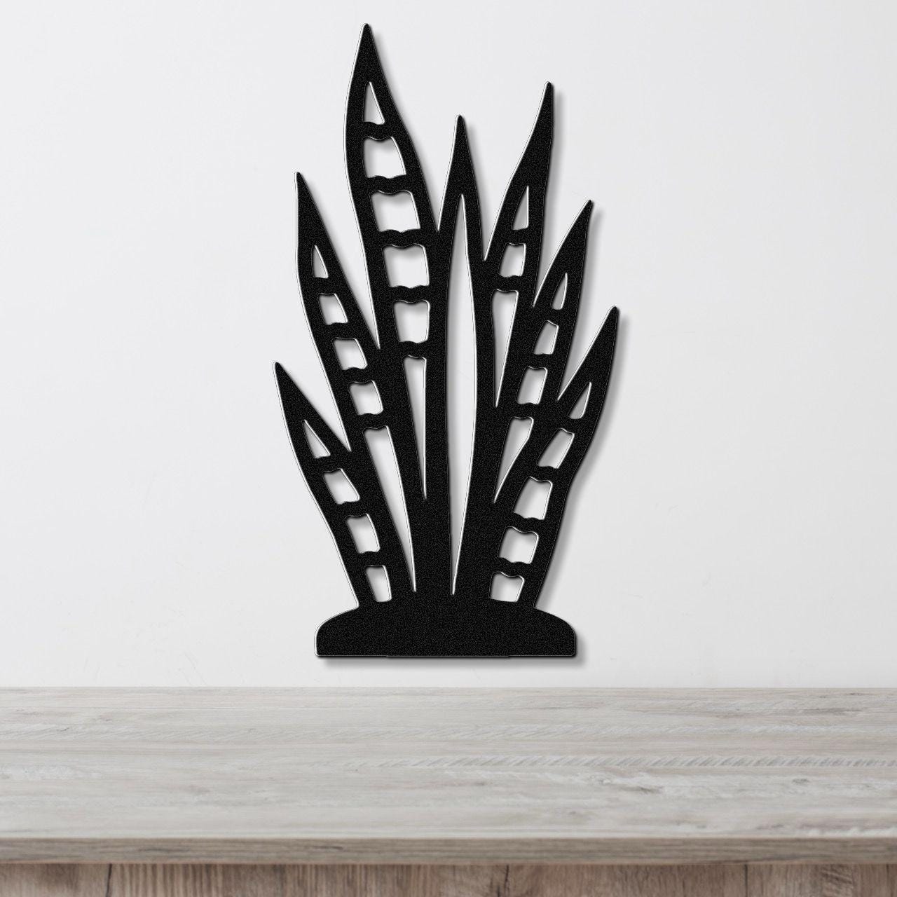 Black metal wall art cutout of a snake plant with leaf-shaped openings, mounted on a white wall above a wooden surface.