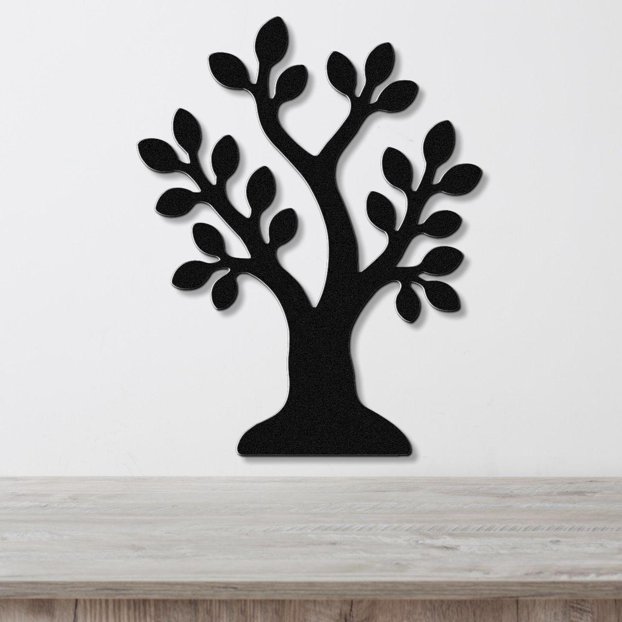 Black metal wall art in the shape of a tree with leaf cutouts, mounted on a white wall above a light wood surface.