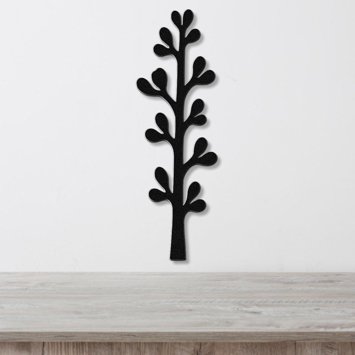 Black metal wall art in a silhouette of an olive branch with leaf cutouts, mounted on a white wall above a light wood surface.