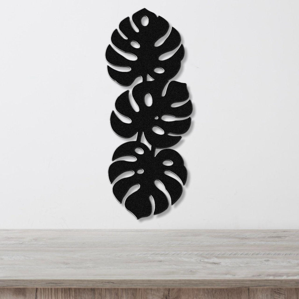Black metal wall art featuring three vertically stacked monstera leaf cutouts, mounted on a white wall above a light wood surface.