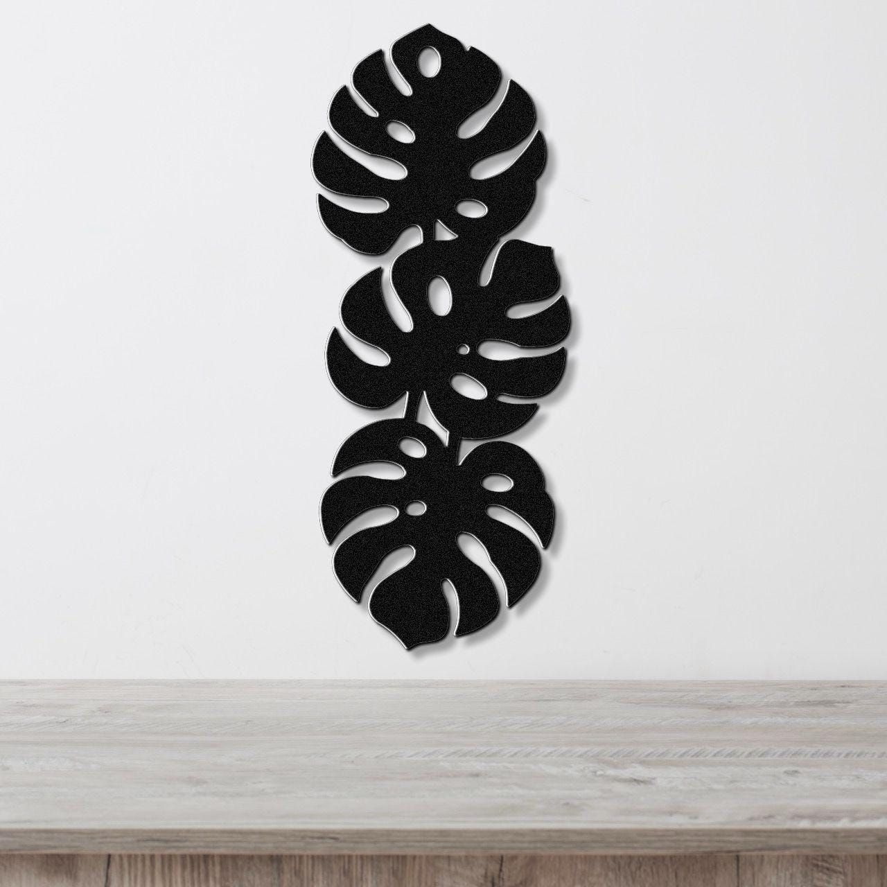 Black metal wall art featuring three vertically stacked monstera leaf cutouts, mounted on a white wall above a light wood surface.
