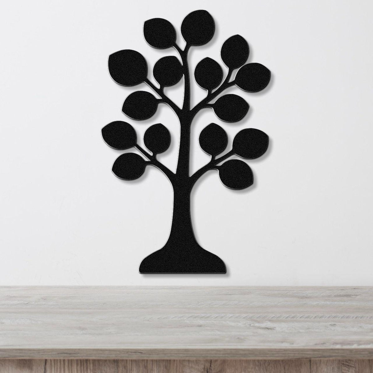 Black metal wall art in the shape of a tree with leaf cutouts, mounted on a white wall above a light wood surface.