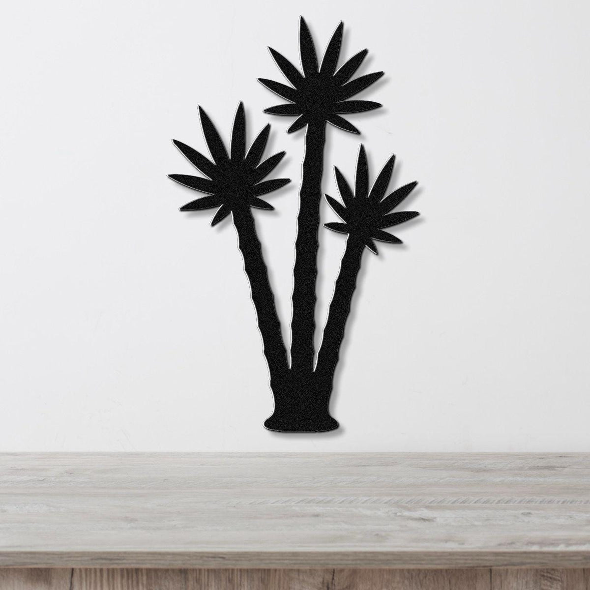 Black metal wall art featuring a silhouette of three palm trees with leaf cutouts, mounted on a white wall above a wooden surface.